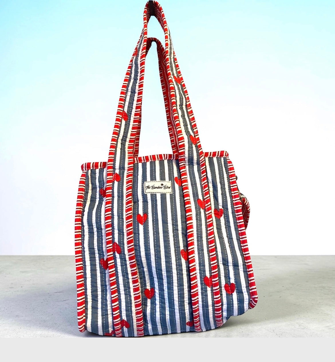 A striped canvas tote bag with red and white vertical stripes, red heart details, and red striped handles, standing on a light gray surface against a white and light blue gradient background.