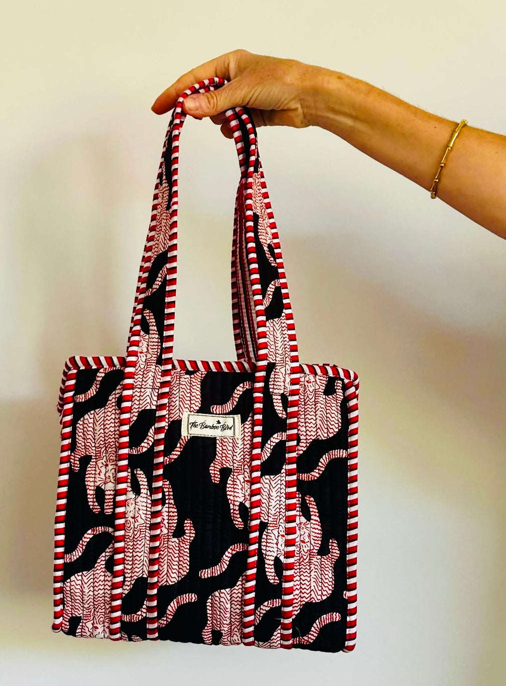 A hand holding a small fabric tote bag with red, white, and black patterned handles and body, featuring a small white brand label that reads 'the textile shop'.