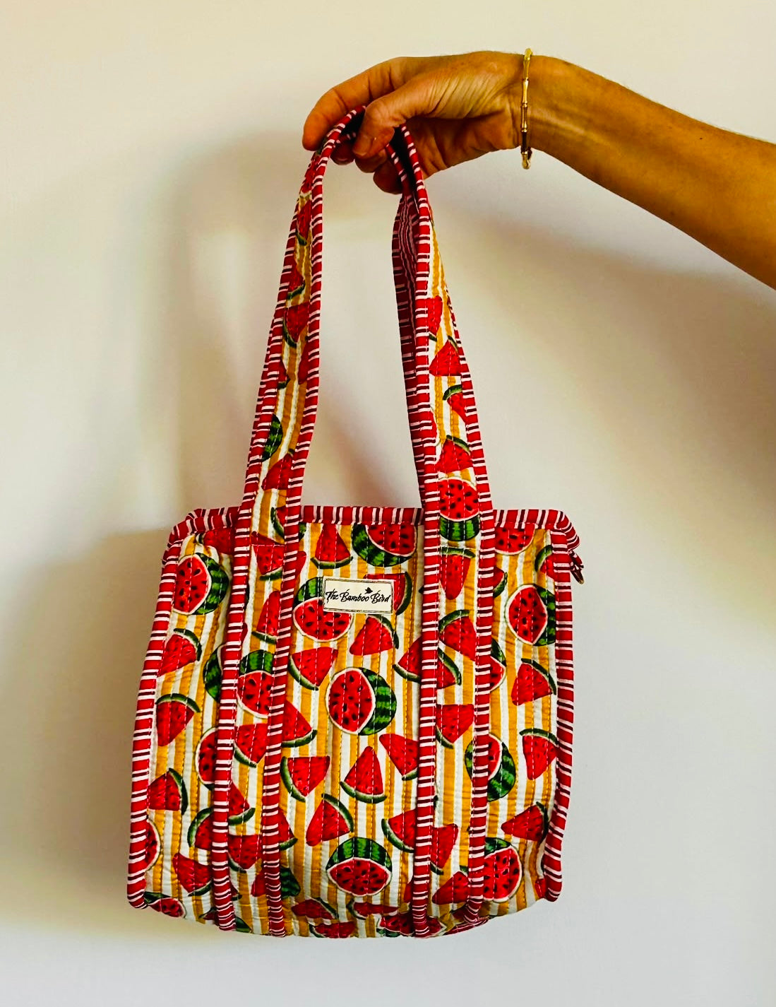 A hand holding a small fabric tote bag with a watermelon print on a cream background, featuring red and green watermelon slices and striped red-and-white handles.