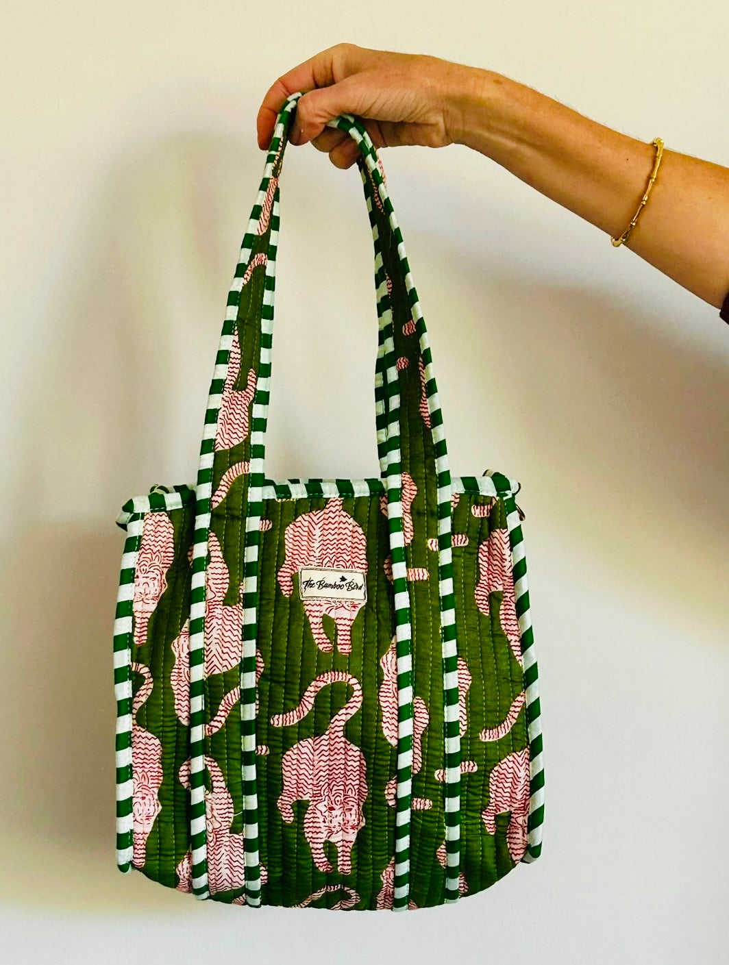 A hand holding a green quilted tote bag with pink elephant prints and green-and-white striped handles, against a plain off-white wall.