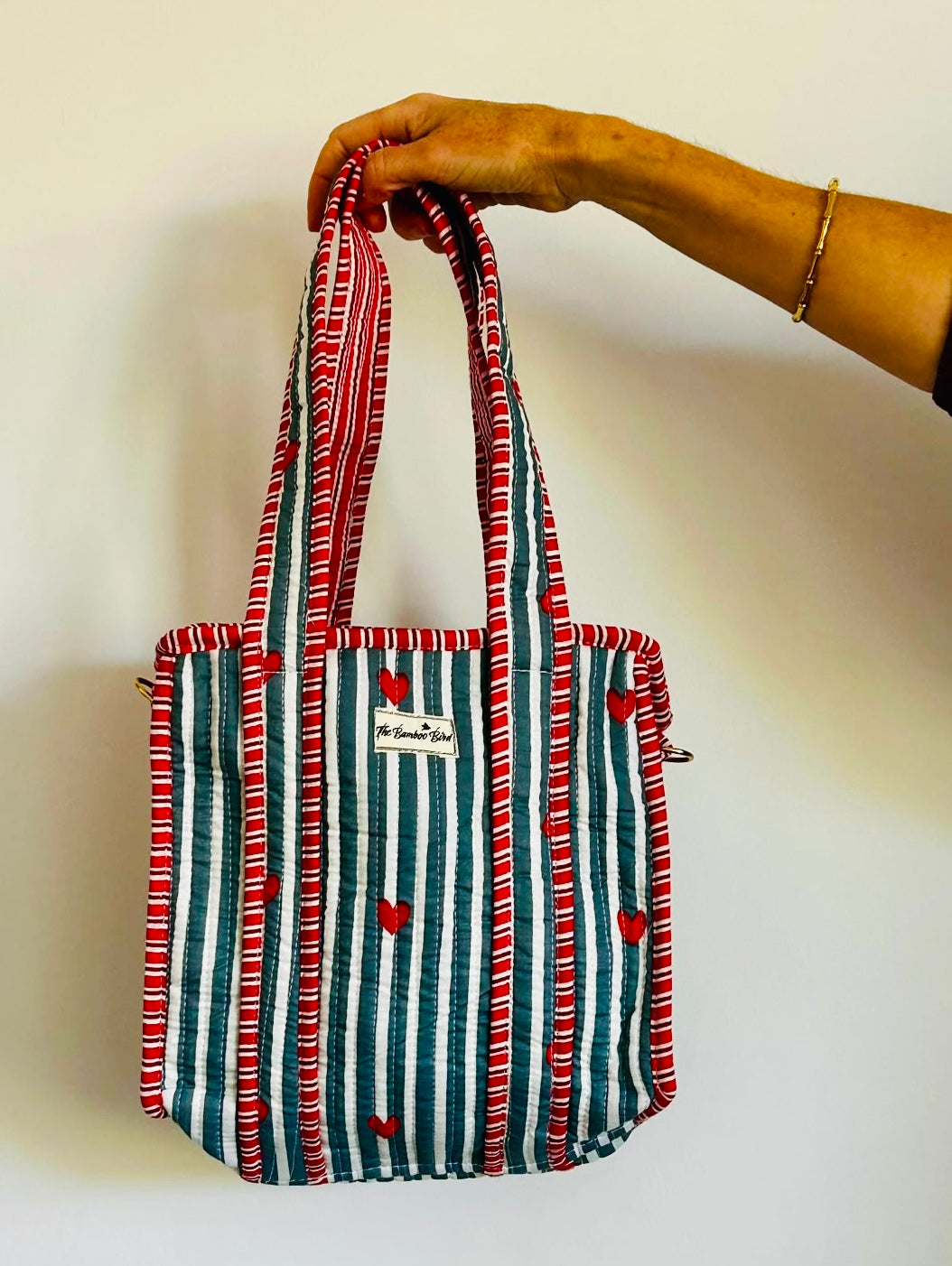 A striped fabric tote bag with red and white wavy borders, vertical teal and white stripes, and small red hearts, held by a person's hand against a plain white wall.