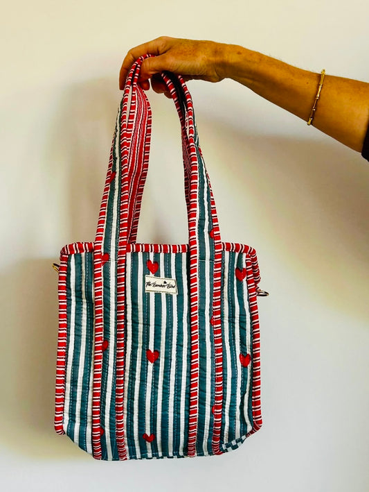 A striped fabric tote bag with red and white wavy borders, vertical teal and white stripes, and small red hearts, held by a person's hand against a plain white wall.