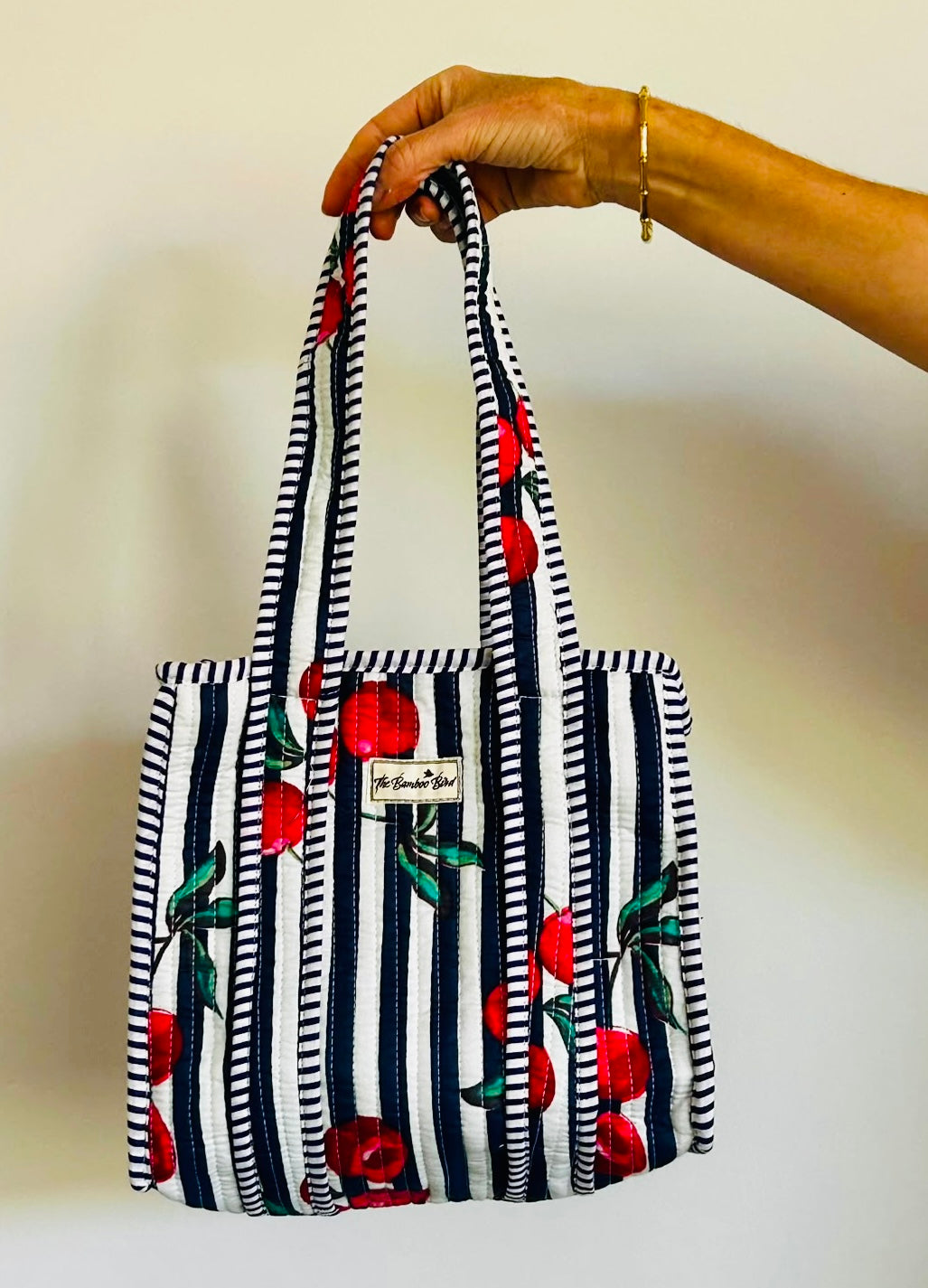 A hand holding a small tote bag with navy and white vertical stripes, red cherries, and green leaves. A small white label is visible on the front.